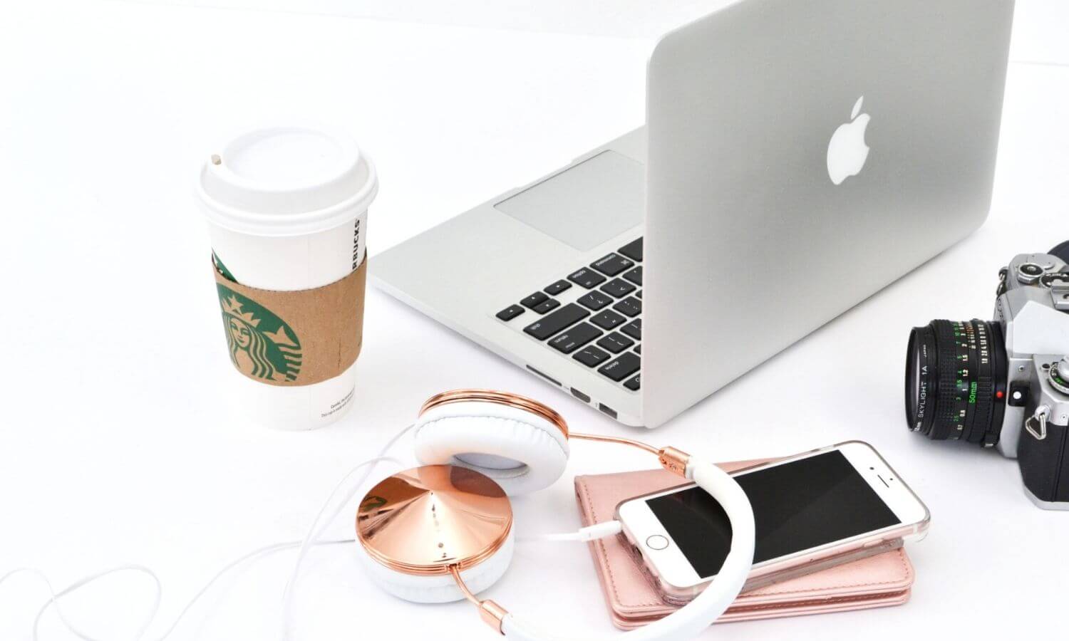 silver mac laptop, rose colored headphones, cell phone, starbucks cup on a white desk