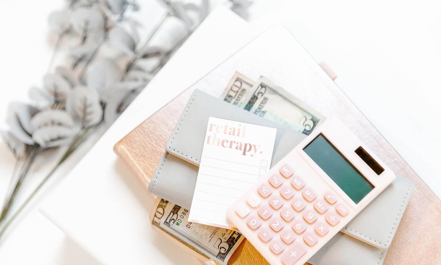 pink calculator, notepad with retail therapy written on it, grey purse, flowers on a white backgound