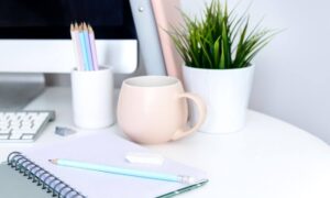 corner of a white desk with notebook laying open with a pencil and eraser on top, monitor , pencils in pencil holder, pink coffee cup and plant in the background