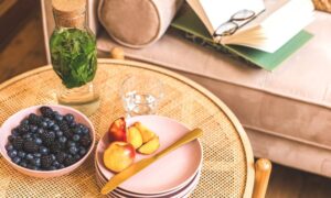 blackberries, blueberries in a white bowl, cut up peaches with a knife on a plate on a coffee table in front of a couch with an open book on it